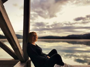 Young woman sitting by sea against sky during sunset