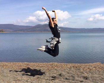 Side view of young woman jumping by lakeshore against sky