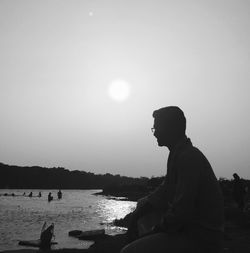 Silhouette man sitting at lakeshore against clear sky on sunny day