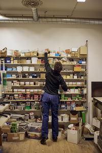 Rear view of mature female worker reaching package on shelf in hardware store