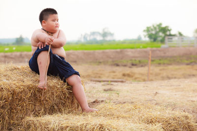 Shirtless boy sitting on hay bale against sky