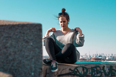 Portrait of young woman sitting on retaining wall against clear sky