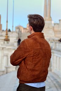 Rear view of man wearing brown jacket while standing on street in city