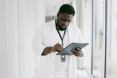 Young male doctor writing on clipboard while standing at hospital corridor