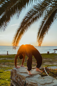 Side view of anonymous male shirtless athlete striking bridge doing yoga poses on rocky fence with empty beach blue sea and palm leafs on background during sunset