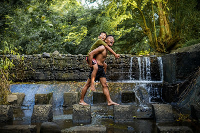 Full length of shirtless man piggybacking son while standing on rock against trees in forest