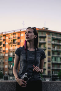 Beautiful young woman holding camera while standing against sky in city