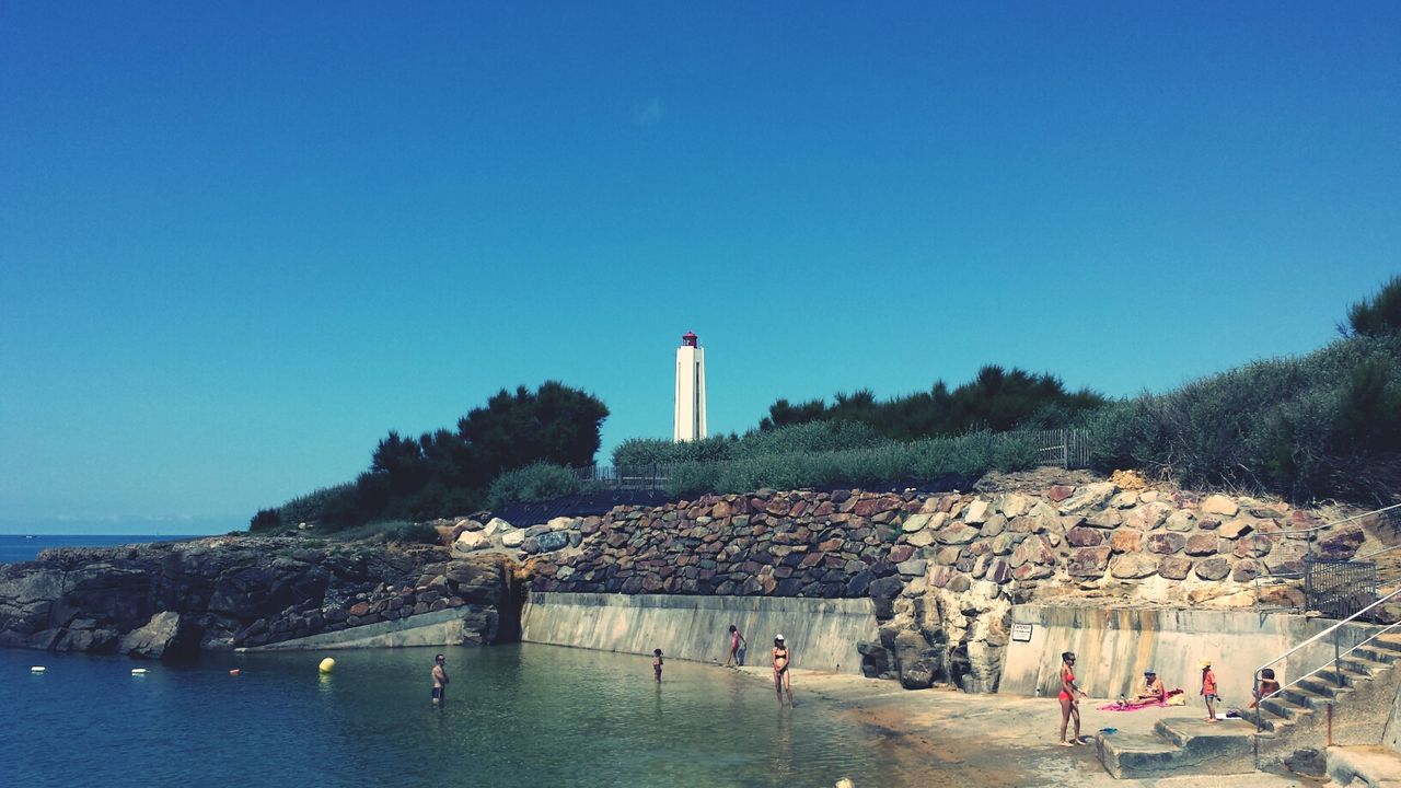 water, clear sky, built structure, architecture, building exterior, copy space, lifestyles, leisure activity, large group of people, men, sea, person, blue, vacations, tourist, rock - object, beach, tourism, mixed age range