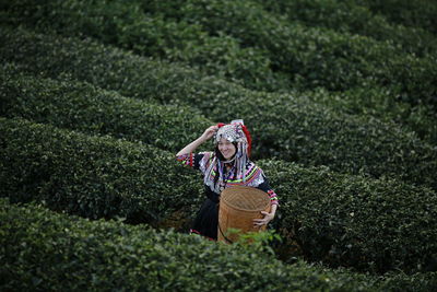 Portrait of smiling woman with wicker basket standing amidst tea crops on farm