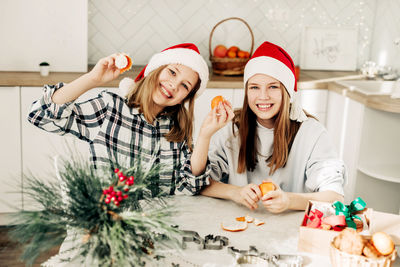 Two cute sisters in a red santa claus hat are sitting in the kitchen and eating tangerines