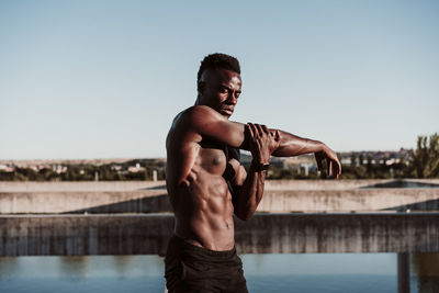 Male athlete doing stretching exercise while standing against clear sky on sunny day