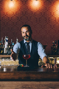 Portrait of smiling mature bartender gesturing by cocktail on bar counter