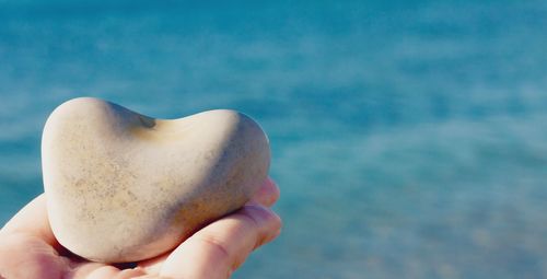 Close-up of hand holding stone in the shape of a heart against blurred background of the blue sea