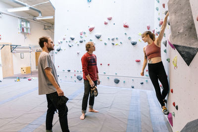 Woman climbing wall while looking at instructor and male student in gym