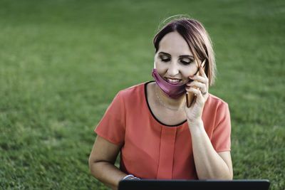 Smiling woman wearing mask talking on phone while using laptop at park