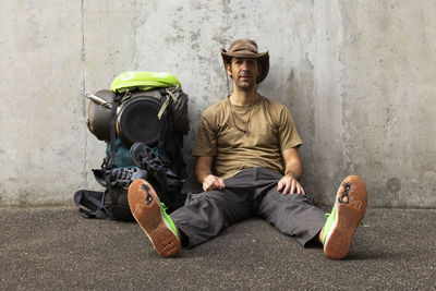 Portrait of man sitting with backpack on footpath against wall