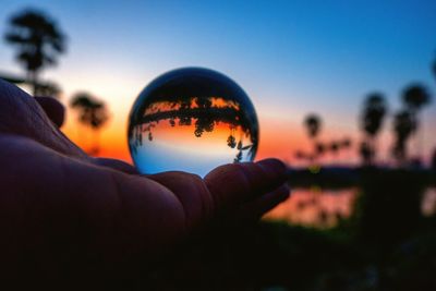 Close-up of hand holding crystal ball against sky during sunset