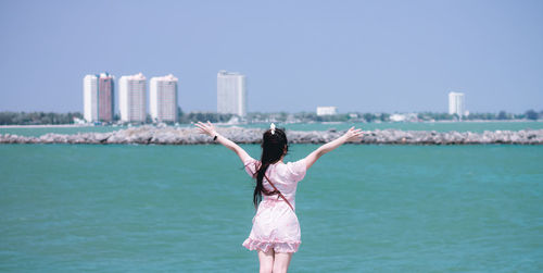Rear view of woman with arms raised standing against sea in city