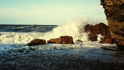 Waves splashing on rocks at shore against sky