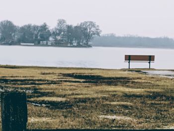 Empty bench on field by lake against sky