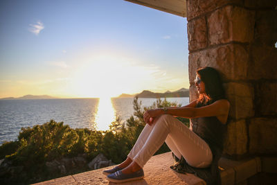 Smiling woman sitting on retaining wall by sea against sky during sunset