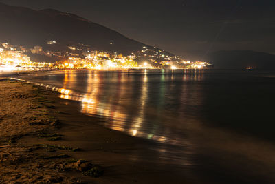 Illuminated city by sea against sky at night