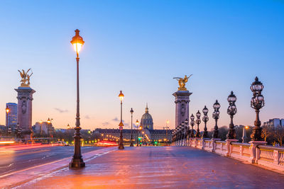 Illuminated street light by building against sky at dusk