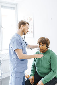 Male medical professional checking pulse of senior patient with stethoscope while standing in examination room