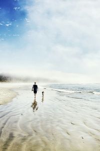 Rear view of man and dog walking on shore at beach against sky