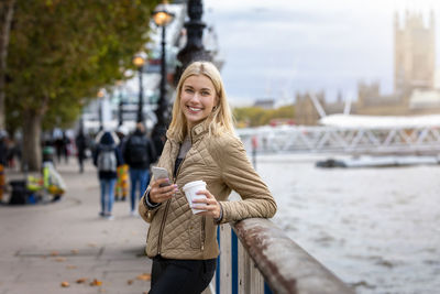 Portrait of smiling young woman holding coffee cup and mobile phone while standing by railing