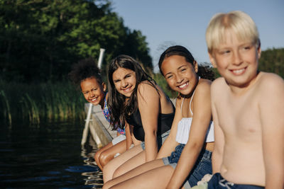 Portrait of happy children sitting together on jetty by lake at summer camp