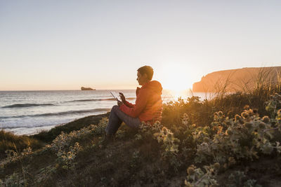 France, bretagne, crozon peninsula, woman sitting at the coast at sunset using tablet