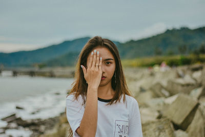 Portrait of young woman hiding face while standing on land against cloudy sky