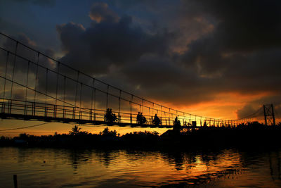 Silhouette bridge over sea against dramatic sky during sunset