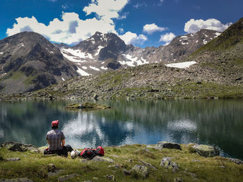 Rear view of man sitting at lakeshore by mountain against sky