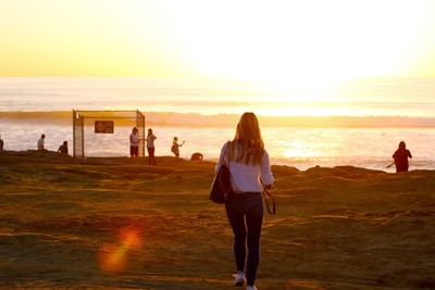 Rear view of woman with shoulder bag walking at beach against sky during sunset