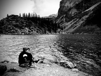 Man sitting on rock by lake against mountain