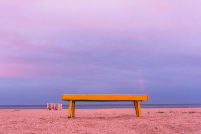 Empty bench on beach against sky during sunset