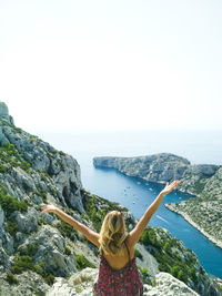 Rear view of woman with arms raised sitting on cliff by sea against sky