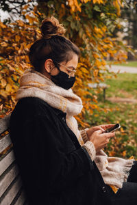 Woman wearing mask using smart phone sitting on bench