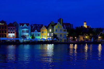 Illuminated buildings by river against blue sky at dusk