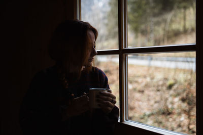 Calm pensive young female enjoying hot tea while sitting near window in dark room of country house in autumn day