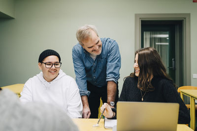 Smiling teacher looking at female student siting by classmate at desk in classroom