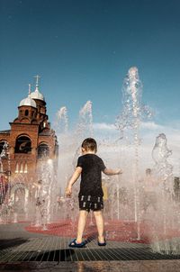 Rear view of boy looking at fountain in city against sky