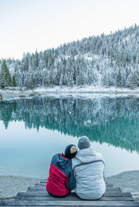 Couple sitting at lakeshore against snowy trees