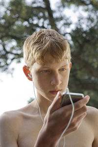 Shirtless teenage boy listening music from mobile phone against tree