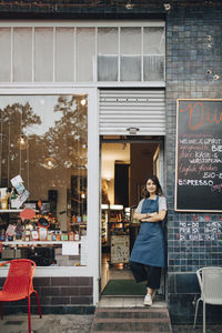 Full length portrait of female owner standing at entrance of deli