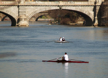 Rear view of people rowing boat in river by bridge