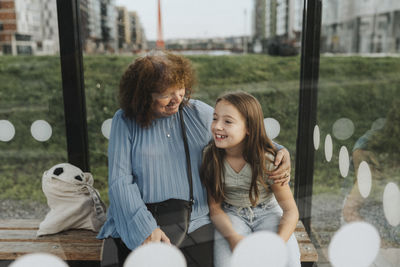 Happy senior woman sitting with arm around granddaughter at bus stop