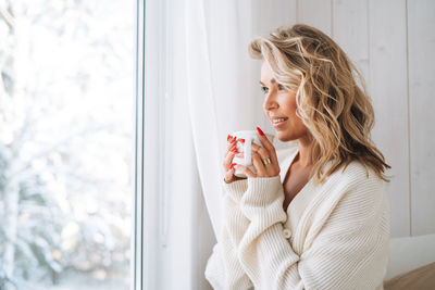 Young woman in white cardigan with cup of cocoa in hands looking at window with winter landscape 